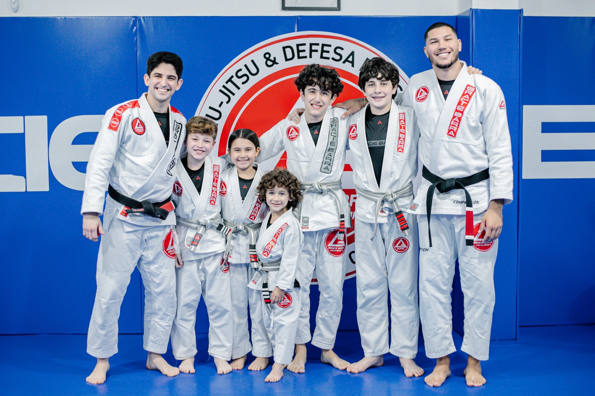 Instrutores e crianças da Gracie Barra Tubarão posando lado a lado diante do escudo da escola