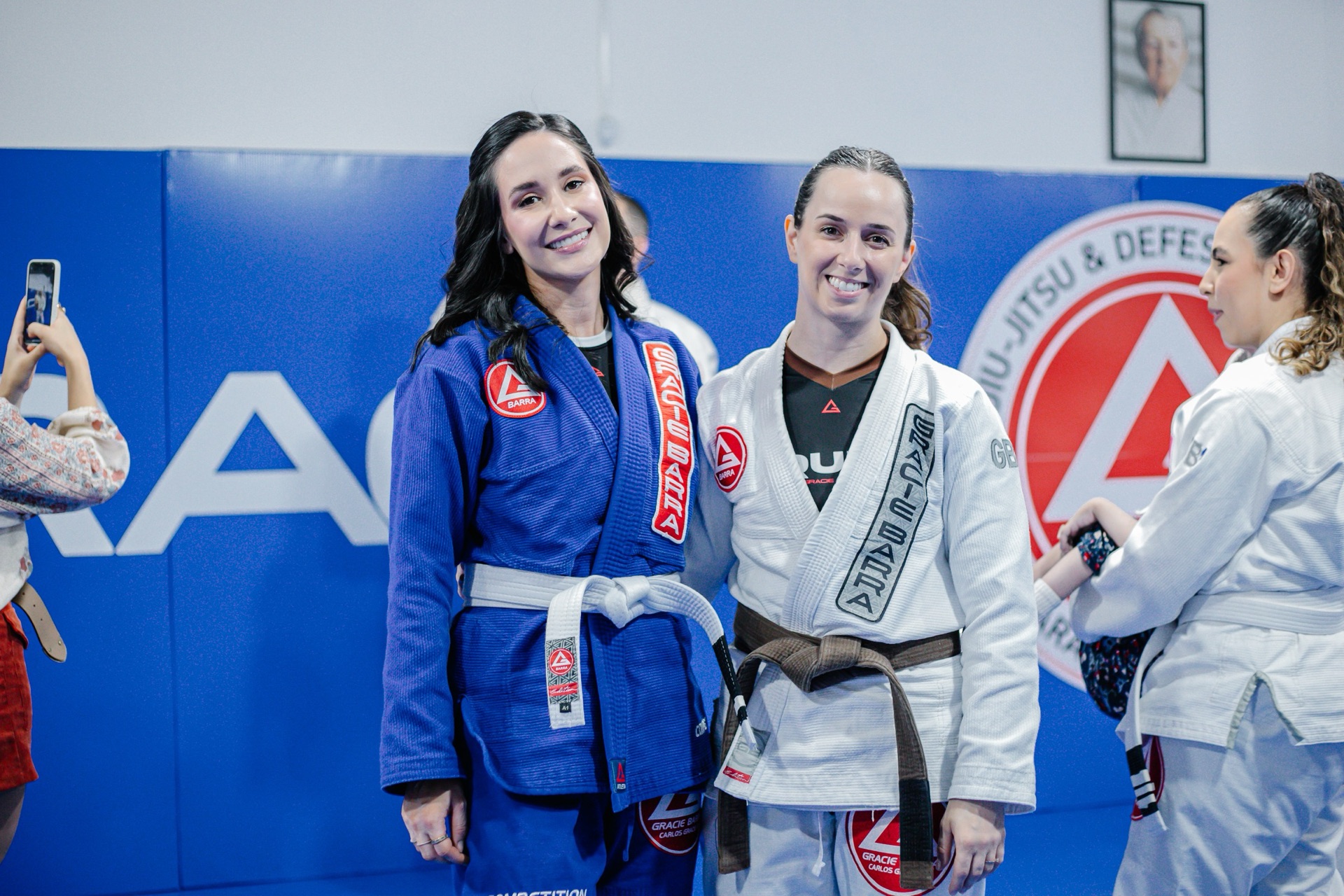 Duas alunas da Gracie Barra Tubarão sorrindo de kimono diante do escudo da escola, uma delas de faixa marrom