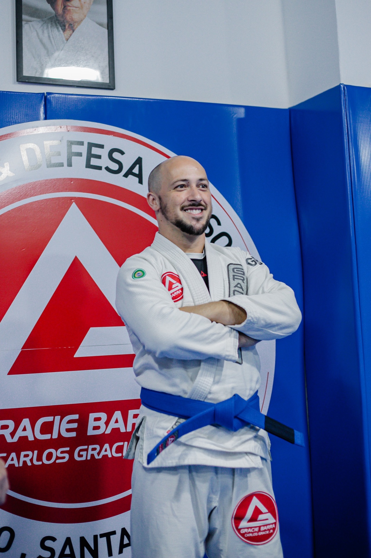 Instrutor de faixa azul sorrindo de braços cruzados diante do escudo Gracie Barra