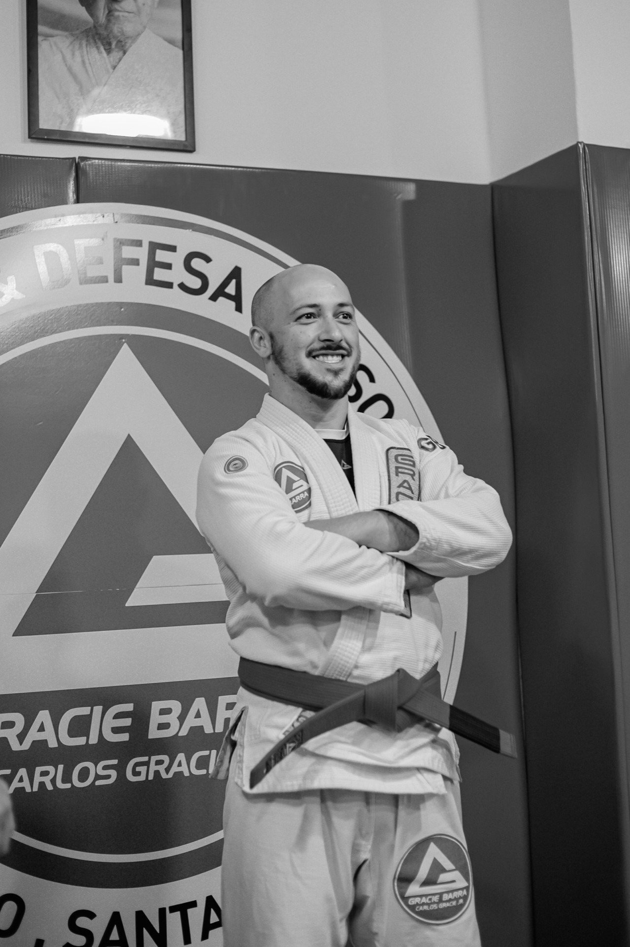 Retrato em preto e branco de instrutor faixa preta de braços cruzados diante do escudo Gracie Barra
