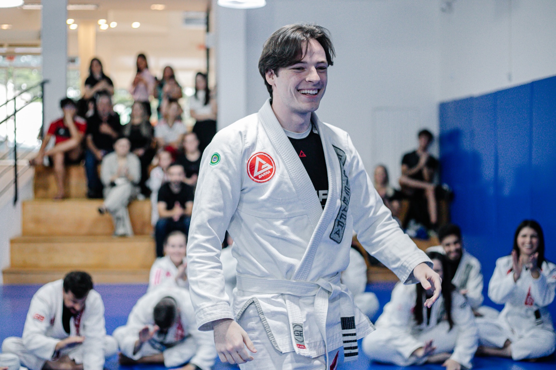 Aluno de faixa branca sorrindo em pé no tatame durante a aula, com colegas de kimono sentados e familiares na arquibancada ao fundo